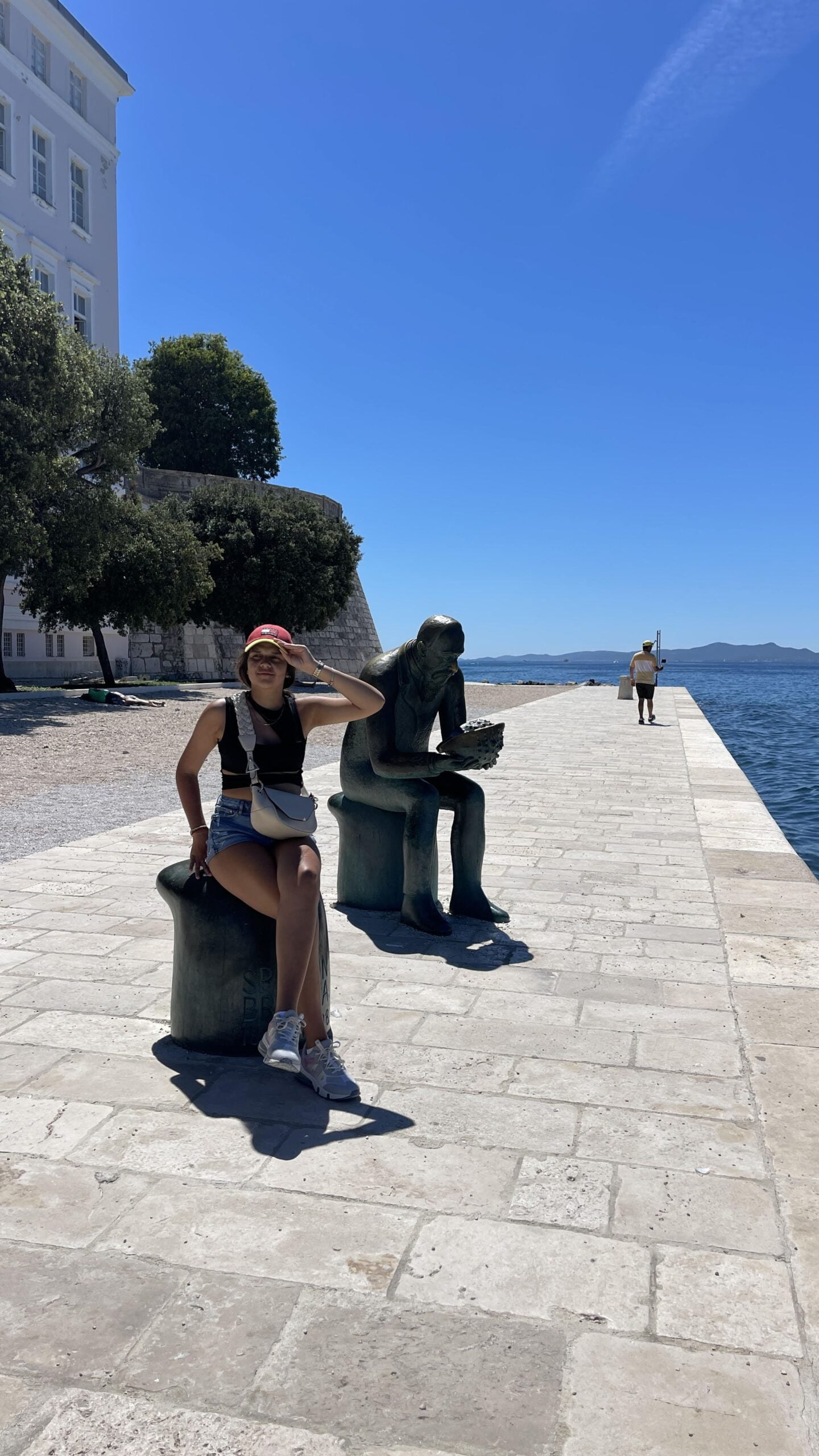 chica sentada al lado de una estatua con un hombre de fondo en Zadar, Croacia